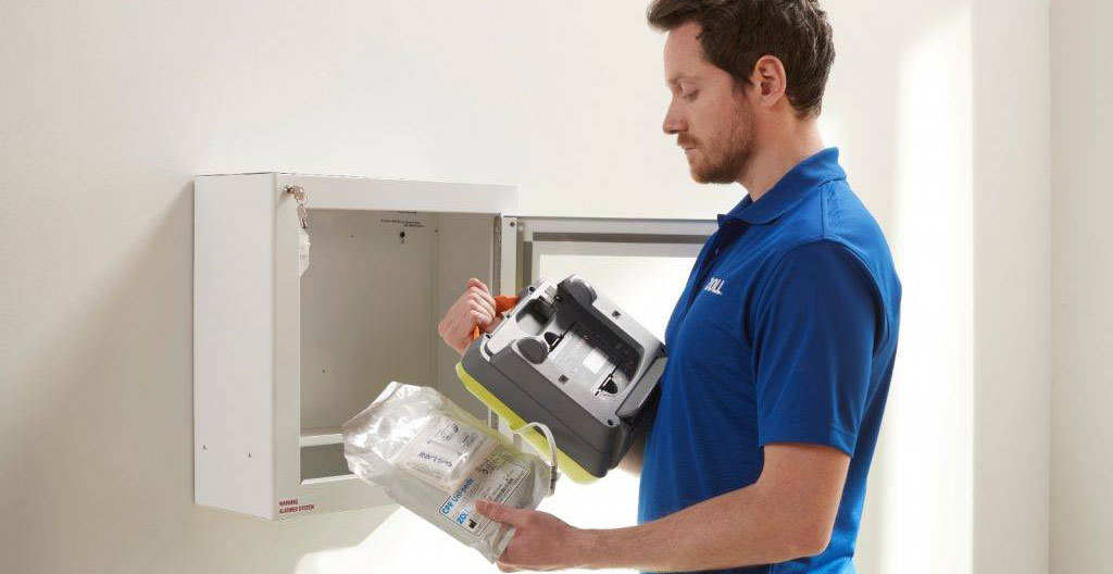 Mains d'un technicien effectuant une vérification de maintenance sur un défibrillateur automatisé externe installé, avec étiquette de maintenance et registre visibles
